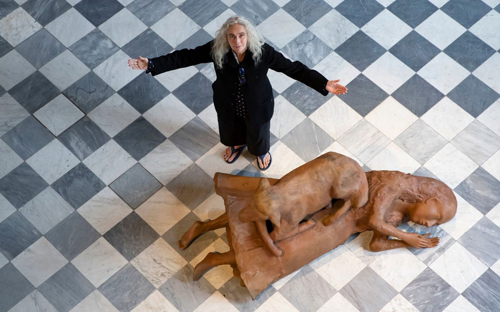 Photograph looking down on Kiki Smith with her arms stretched open. She stands beside Rest Upon, a sculpture of a girl or young woman laying on her side with a sheep laying on top her torso and legs