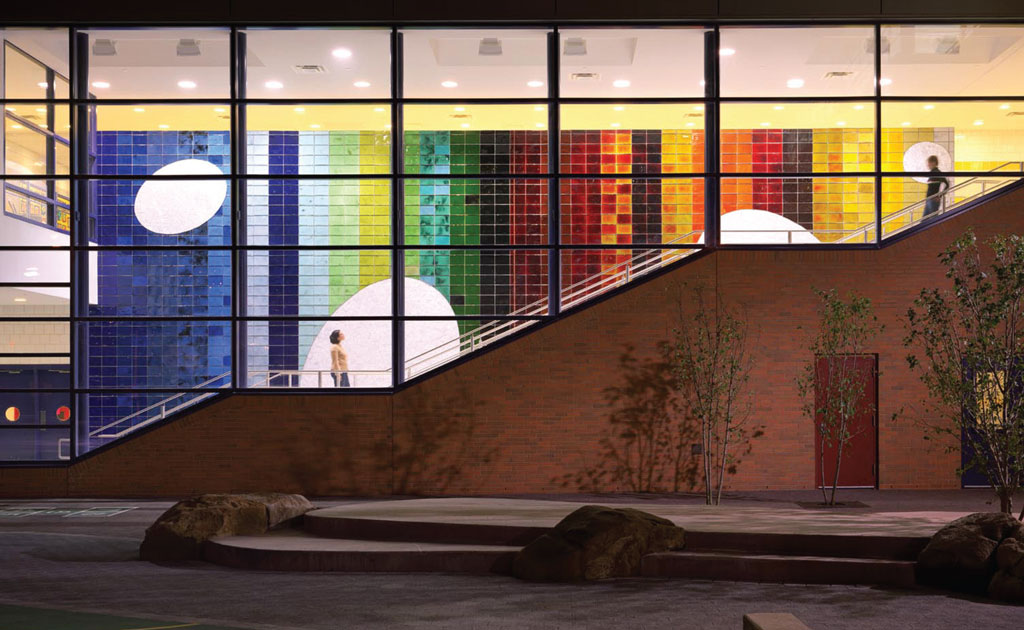Photograph showing the collaboration between architect Paul Broches and artist Ned Smyth. Shot outside during low light, the lit interior of the building highlights a staircase that rises in front of a colorful artwork that spans its length