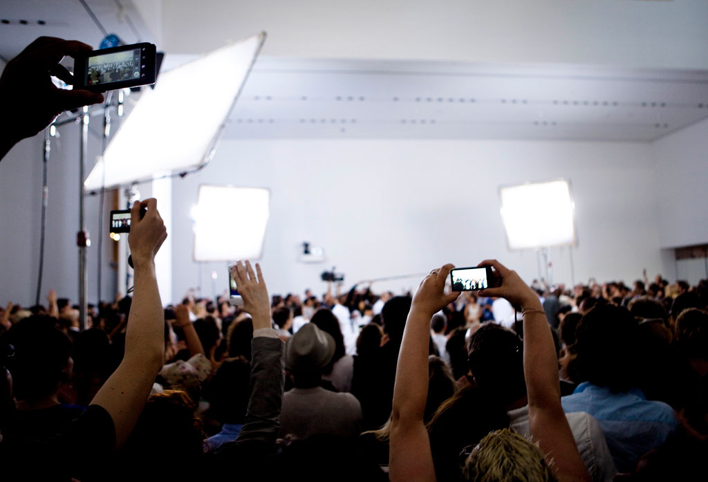 Museum visitors crowd the brightly-lit performance space. Many hold smartphones above their heads, capturing the performance of The Artist Is Present. Marina Abramovic is just barely visible within the throng of people.