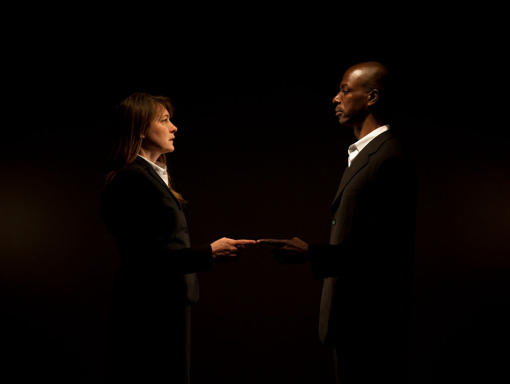A dramatically-lit photo showing two artists dressed in dark suits who stand close together, making eye contact, with index fingers extended, but not touching.