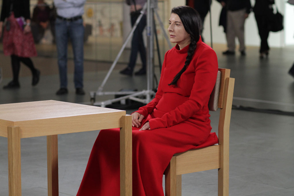 A view of the performance of The Artist Is Present. Marina Abramovic, wearing a long, flowing, red gown, sits across a table, with a crowd of viewers behind her.
