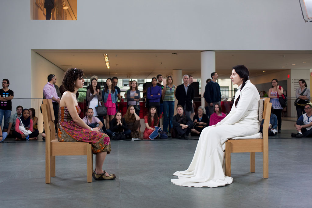 A view of the performance of The Artist Is Present. Marina Abramovic, wearing a long, flowing, white gown, sits a few feet across from a visitor, with a crowd of visitors watching behind them.