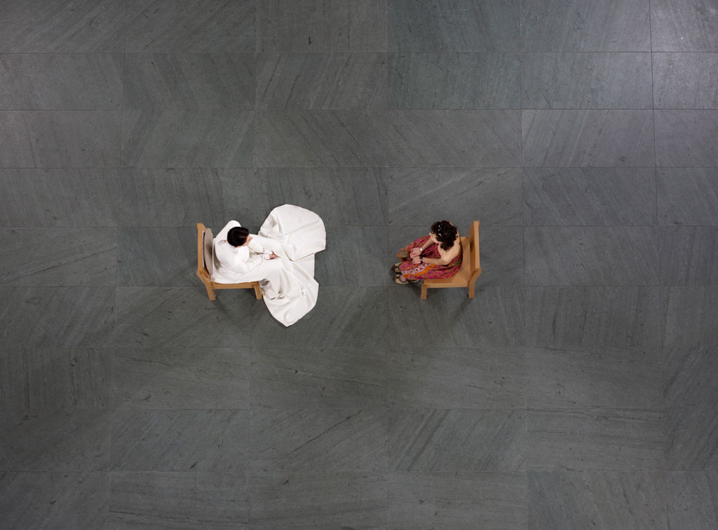 An austere bird's-eye view of the performance of The Artist Is Present. Marina Abramovic, wearing a long, flowing, white gown, sits a few feet across from a visitor.