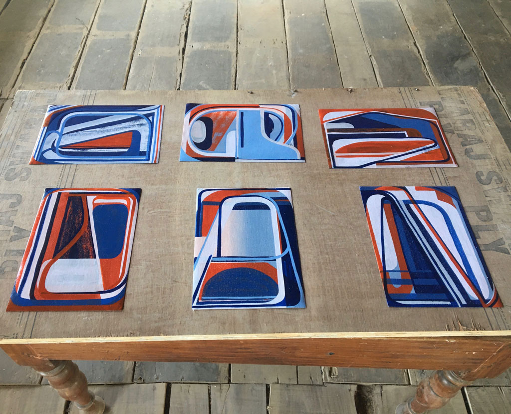 A birds eye view of six small abstract paintings by Tom Buckhardt lined up on a table. Each features a variation on the red and blue color scheme