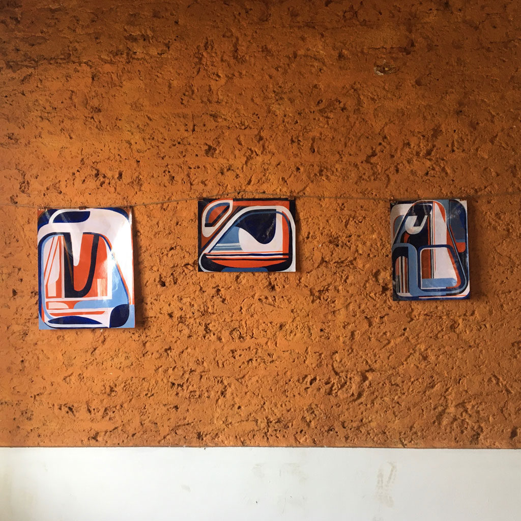 Three small abstract paintings by Tom Buckhardt hang from wire on a rugged wall. Each features a variation on the red and blue color scheme