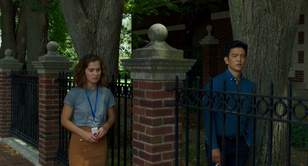 Casey and Jin walk in contemplation beside each other on opposite sides of a wrought, brick, and concrete fence designed in a traditional style.