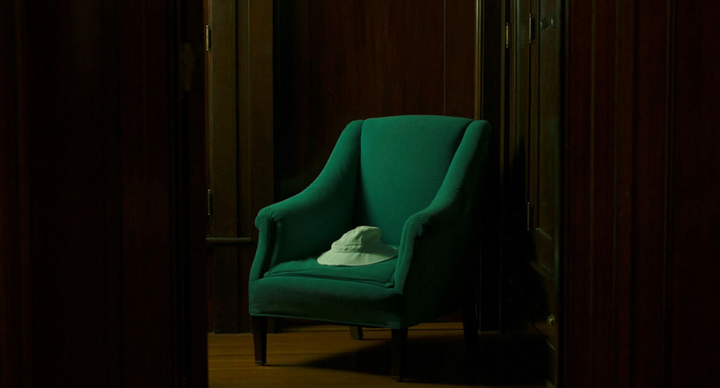 This moody shot shows a lonely, floppy green hat sitting on a darker green arm chair at the end of a hallway framed in dark wooden panels.