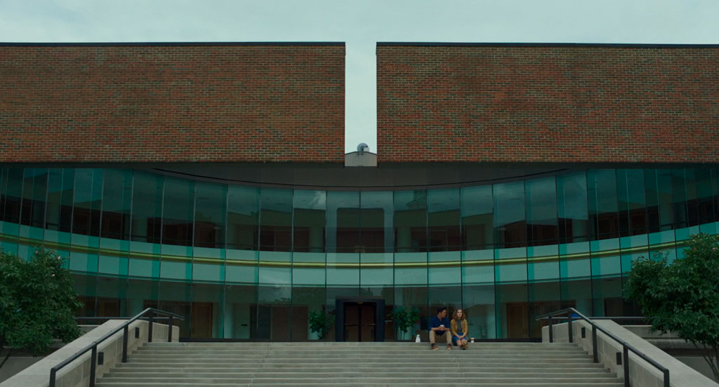 This long shot shows Jin and Casey sitting at the top of the staircase that leads to Columbus City Hall, designed by Edward Charles Bassett. The building features a wall of rounded, transparent, floor-to-ceiling glass, framed by two brick, cantilevered arms. A clear sky peeks through the space between the two arms.