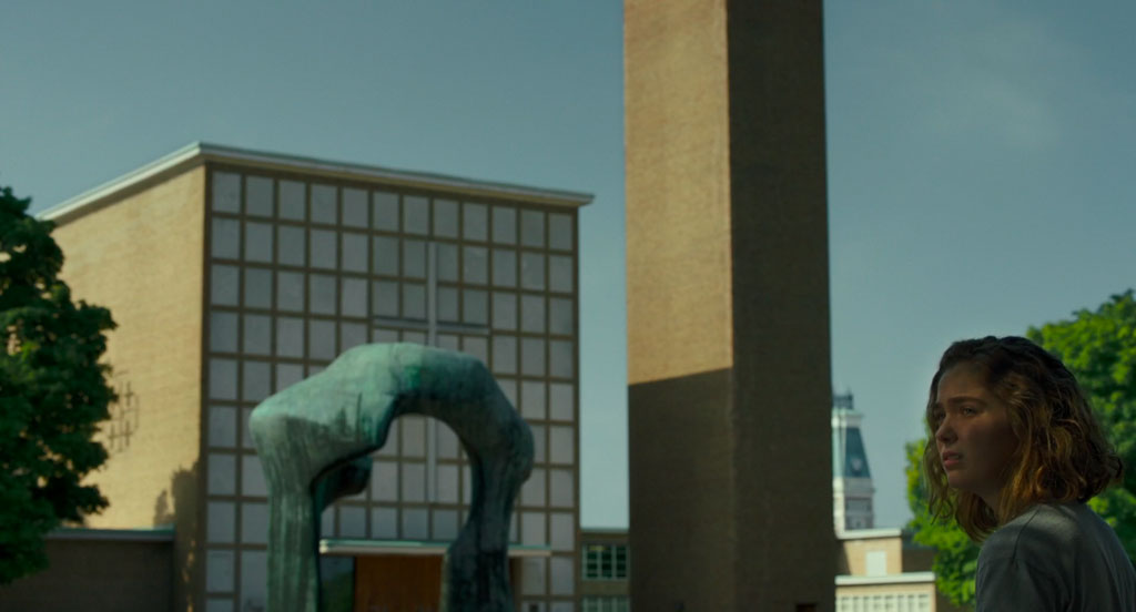 Casey looks across the plaza outside the Cleo Rogers Memorial Library. In the background we see a Modern, patinated bronze sculpture by Henry Moore entitled Large Arch. Further in the background is the First Christian Church and its clocktower, designed by Eliel Saarinen. We see the building’s simple, geometric design, its large cross, and its buff brick and limestone materials.