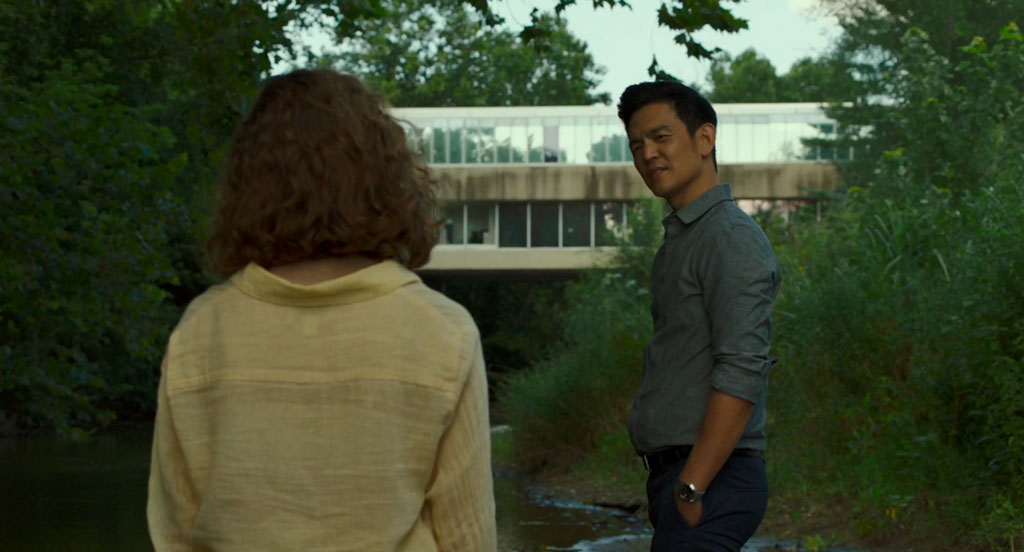 Jin and Casey pause to talk on a path alongside Haw Creek. In the background is a glimpse of the building James Stewart Polshek designed for the Columbus Regional Health Hospital Mental Health Services facility. We see its glass and concrete bridge that crosses above the river, connecting the main hospital building with the Mental Health Services facility.