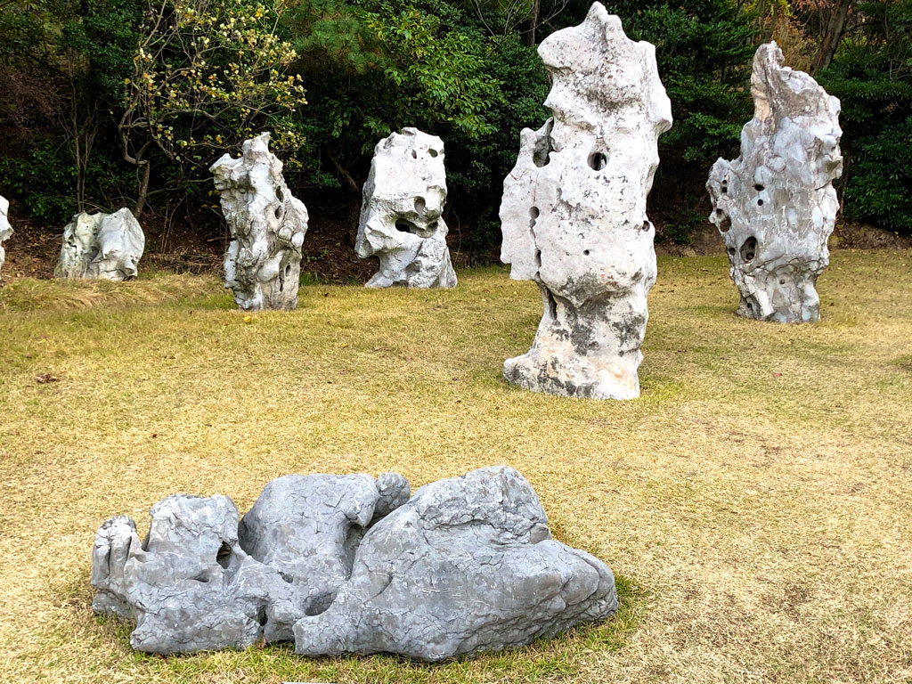 This photograph shows an arrangement of large stones on a lawn. The stones have a natural appearance, with nonuniform surfaces displaying indentations and holes, almost as if worn over time by the sea or wind. Their standing arrangement gives them a humanlike presence.