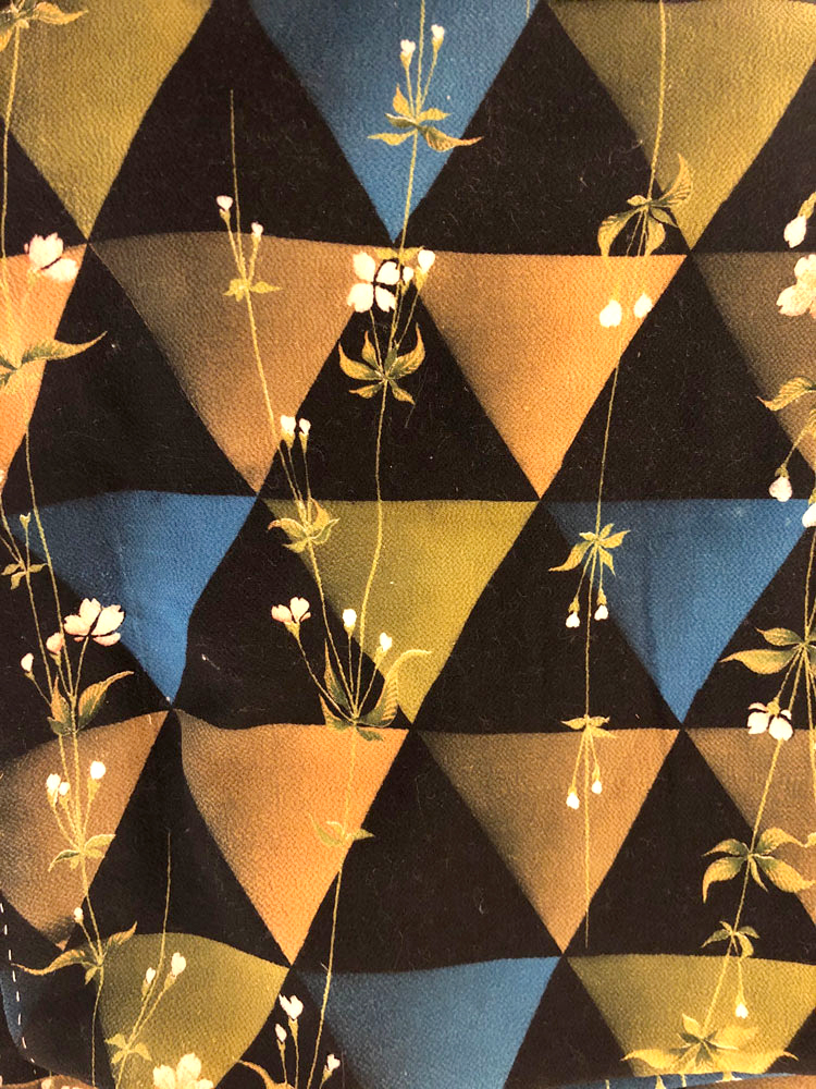 This detail shot shows a well-preserved antique kimono’s quiet, yet striking pattern made up of repeating triangles overlaid with a few stems of delicate, flowering grasses. The triangle pattern is dominated by a dark background, with alternating golds, oranges, and blues that appear to glow, as if lit from within.