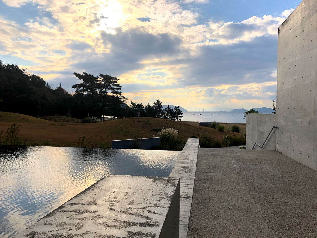 This dreamlike photograph features a concrete building and walkway positioned alongside the flat surface of a pool. Beyond, the view stretches through rolling lawns, forests, and out into a bay, with mountains and a cloudy sky beyond.