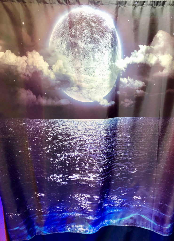 This photograph shows a curtain with a glowing full moon rising among clouds and stars over a peaceful ocean.