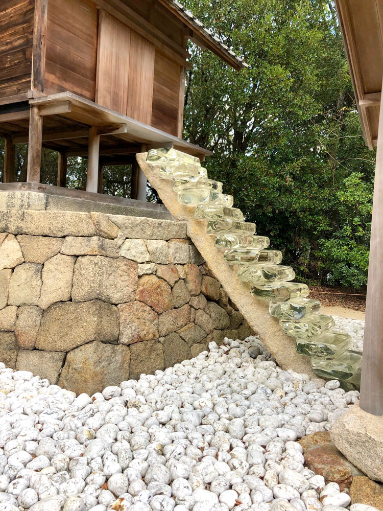This shot shows a staircase made of large chunks of glass leading up a stone foundation and wall to the door of a squat, wooden tower. The ground surrounding the structure is covered with contrasting, lighter-colored rocks.