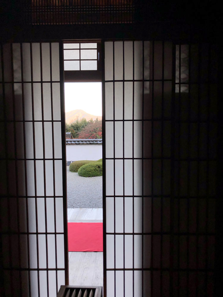 This photograph shows a view of a garden framed by beautiful rice paper doors and a dark interior room. The garden contains gravel; shaped, low bushes; and a simple retaining wall. In the distance are trees with autumnal colors, mountains, and a bright sky.