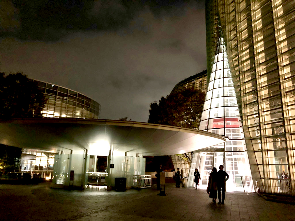 This nighttime shot shows an outdoor plaza, silhouettes of people walking, and a cluster of unusual buildings lit against the dark, cloudy sky. The foremost building is designed as a low, round shape topped by a wider disc. Another is a series of panes of glass forming a transparent, triangular entrance to a larger building.