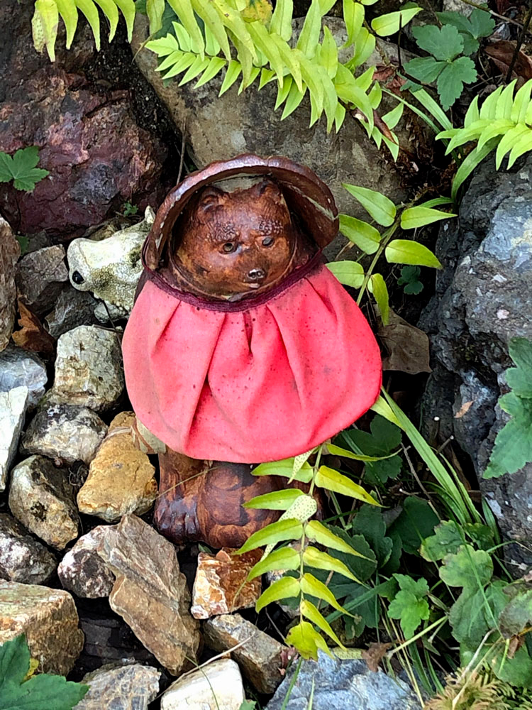 This photograph focuses on a small statute of an animal draped in a bright red cloth. The statue sits outside among rocks and ferns.