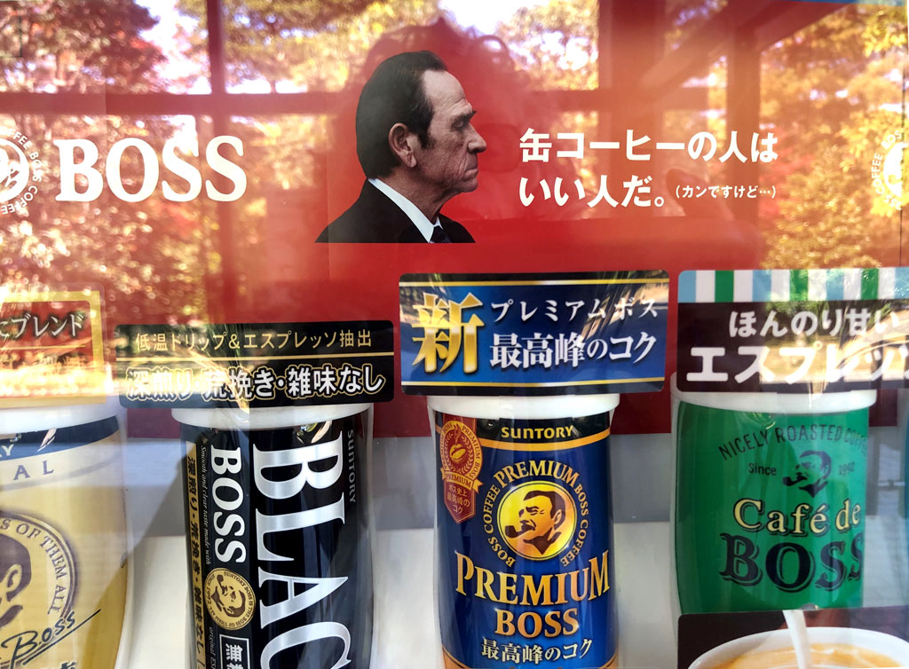 This close-up shot of a vending machine showcases a range of coffee drinks available for purchase. A banner across the machine’s glass contains the Boss coffee branding, including the distinguished profile of actor Tommy Lee Jones, dressed in a suit or tuxedo.