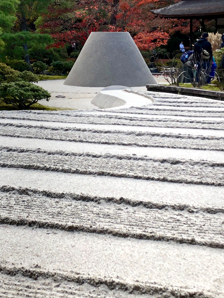 Shot from a low angle, looking up, this photograph shows alternating waves of combed gravel leading up to a mound. Beyond this beautiful, monochrome display are vibrant bushes and trees with fall colors.