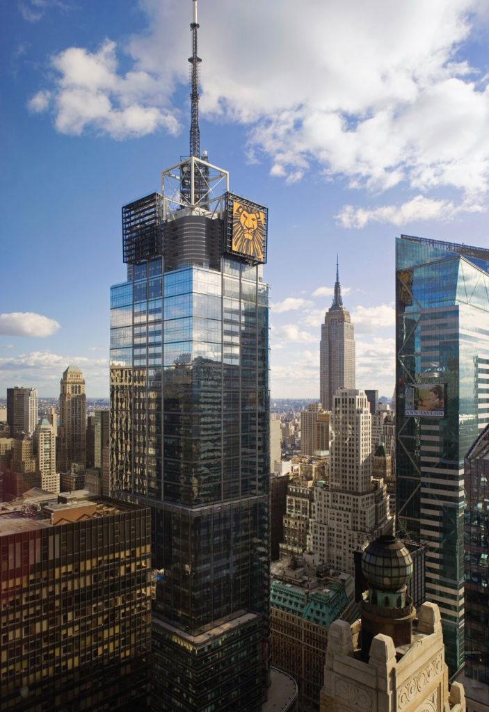 This aerial photograph shows the upper floors and spire of 4 Times Square stretching into the sky.  The glass tower sits among other New York City icons including Hearst Tower and the Empire State Building.