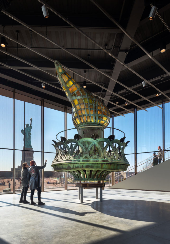 Visitors to the Statue of Liberty Museum observe a display of the statue’s flaming torch.  This room contains floor-to-ceiling glass windows with views of the statue, exterior plaza, and the New York Harbor.