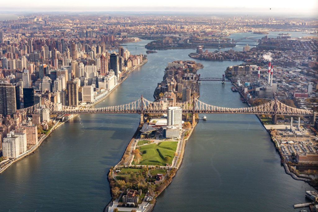A birds eye view of Roosevelt Island in New York City, flanked by Midtown Manhattan’s skyscrapers, the industrial waterfront of Long Island City, and the Queensboro Bridge stretching across the East River to connect all three.  At the center rises the House at Cornell Tech, a 26-story concrete and glass building that follows a seemingly similar design to the area’s older, non-Passive House buildings.