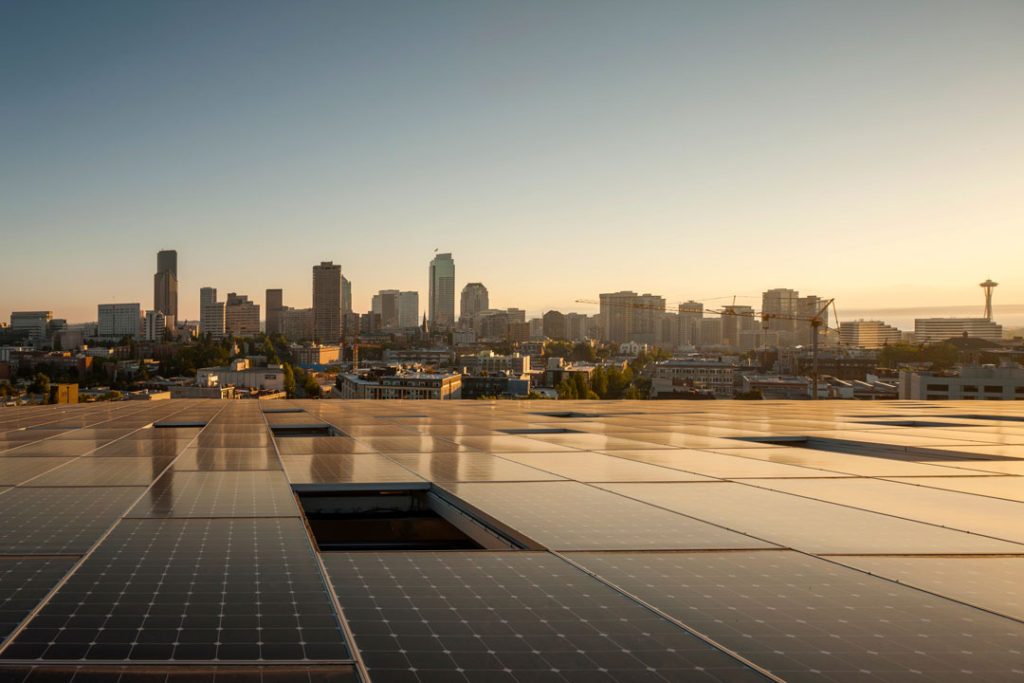 This photo shows the rooftop of the Bullitt Center covered in solar panels.  Beyond the building is the Seattle skyline, including the iconic Space Needle, and a large construction crane.