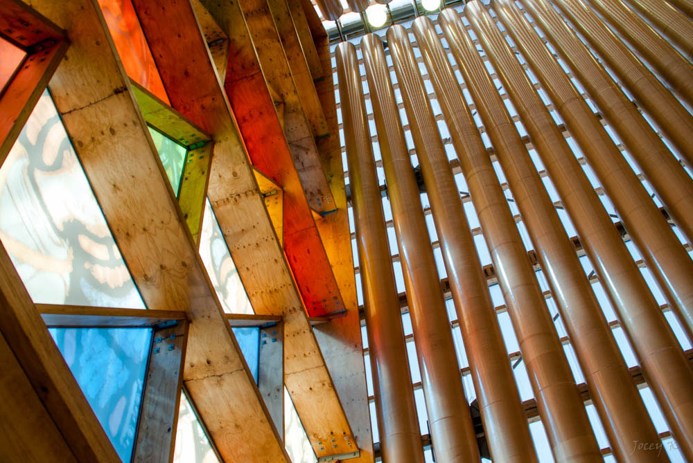 A beautiful detail of the Cardboard Cathedral showing the builing's simple, unique, and climate change-friendly construction.  Daylight shines through pillars of cardboard and multicolored triangular panes of glass.