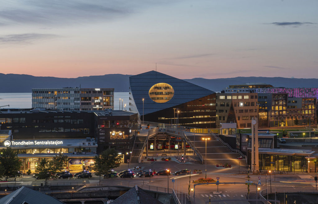 A twilight photograph of the exterior of Powerhouse Brattørkaia building showing its central location near other commercial buildings, a light rail station, and the Trondheim waterfront. The building’s unique design includes a sloped roof with a circular cut-out revealing warm lighting and windows within the interior.