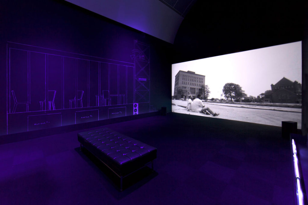 A photograph of a room at the Museum of Contemporary Art Chicago with a bench and projector displaying Afterword via Fantasia by Catherine Sullivan, created in collaboration with George Lewis and Sean Griffin. On the far wall is a drawing by Charles Gaines that shows the outlines of chairs and desks on a raised platform, stage, or blueprint for a building. The video shows a black and white image of two men sitting on a lawn with buildings and mature trees in the distance.