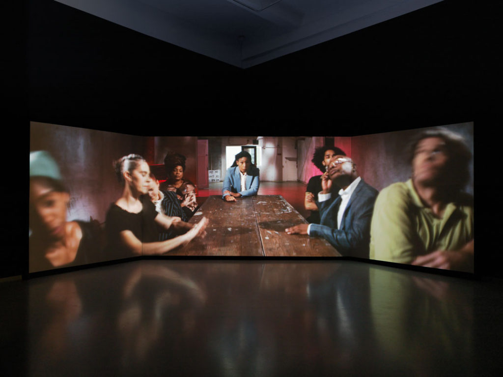 A photograph of The Startled Faction by Catherine Sullivan. The video work spreads across three large screens. This still shows a group of people sitting around a long table, each with their own concerns, some looking at the ceiling, some facing the camera, some looking at their hands. At the head of the table is a Black woman in a suit looking head on at the camera with a look of surprise or mockery.