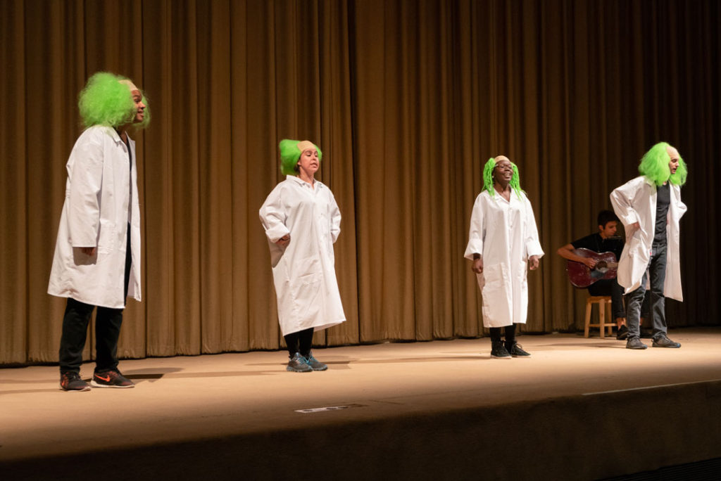 A photograph of Misses Gaines searches out her darky, Cato, by Pope.L. The scene shows four performers on stage in white medical coats and new green wigs, singing and dancing. At the back of the stage, a guitarist plays while sitting on a stool.