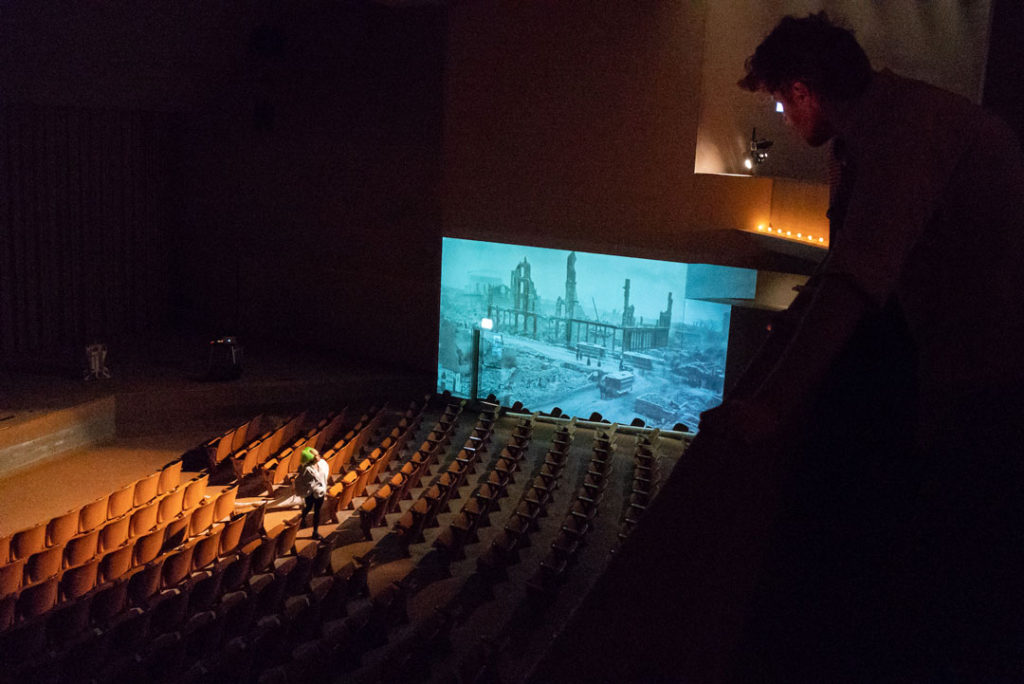 A photograph of a performance by Pope.L entitled Misses Gaines searches out her darky, Cato. The scene shows an observer looking down at a theatre from its balcony. An actor in a medical coat and neon green wig performs in the aisle; the theatre’s seats are empty. Offstage and beyond the rows of seats is a large projection of a historic city in ruins, possibly after a bombing. Despite the decay in the scene, the street is busy with street cars, horse-drawn vehicles, and people walking.