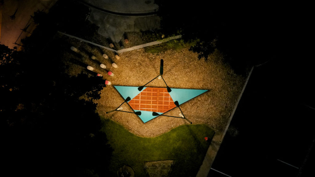 A birds eye view of Jessica Stockholder’s Save on select landscape & outdoor lighting: Song to mind uncouples. The work is shown here at night, with six illuminated lamps focused on a triangular platform below. At the edge of the circle of light are concrete bollards of different sizes, shapes, and colors, some with splashes of hot pink paint across their heads.