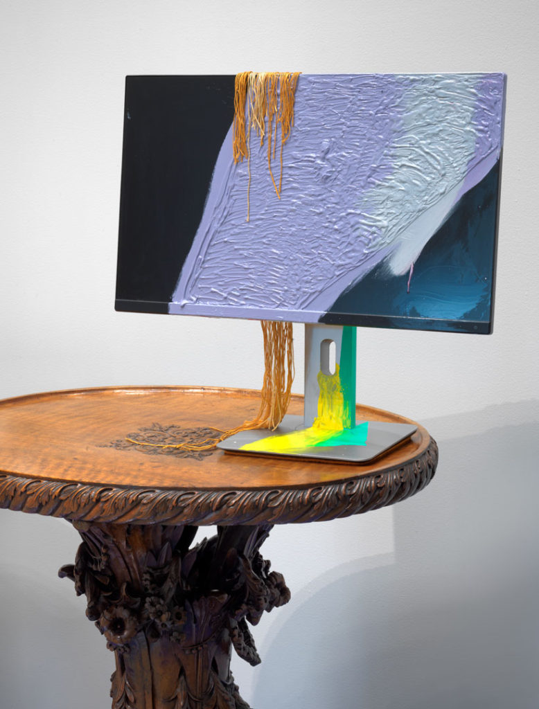 A sculpture by Jessica Stockholder made from a computer monitor that has been painted across its screen and base and draped with thread. The artwork is displayed on a carved, wooden antique table.