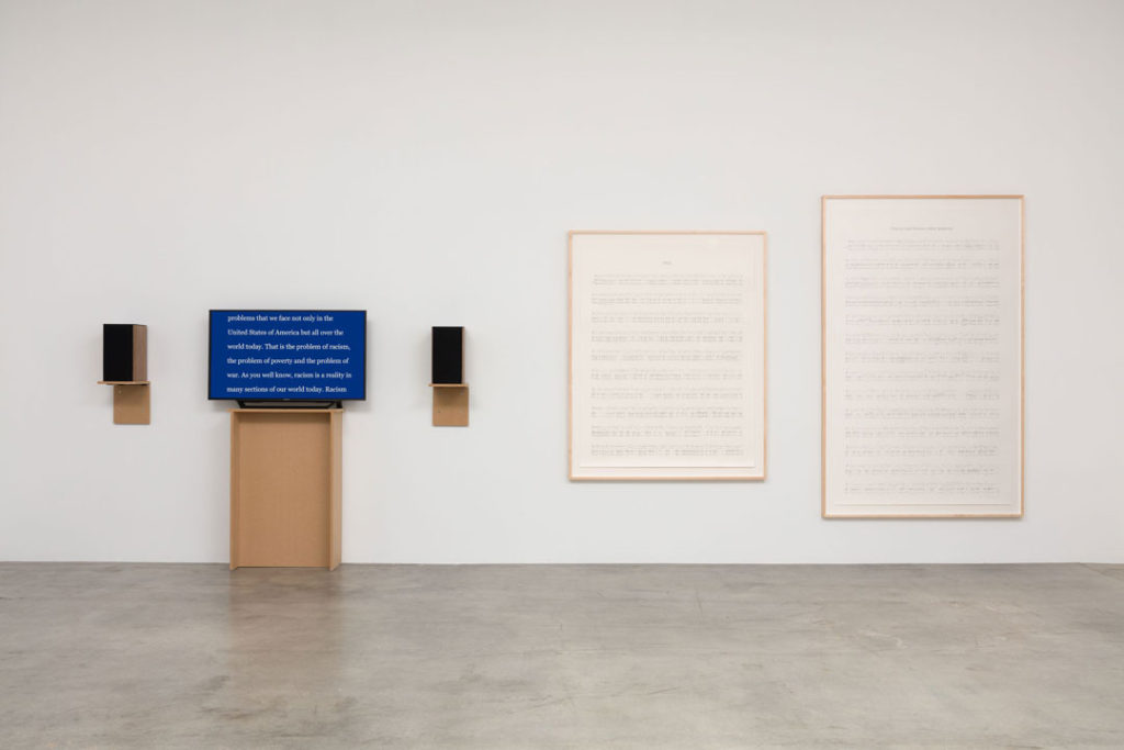An installation view of Manifestos 3 by Charles Gaines.  Along a gallery wall we see a monitor displaying lines of text and two mounted speakers.  Beside these are two large framed drawings of sheet music.