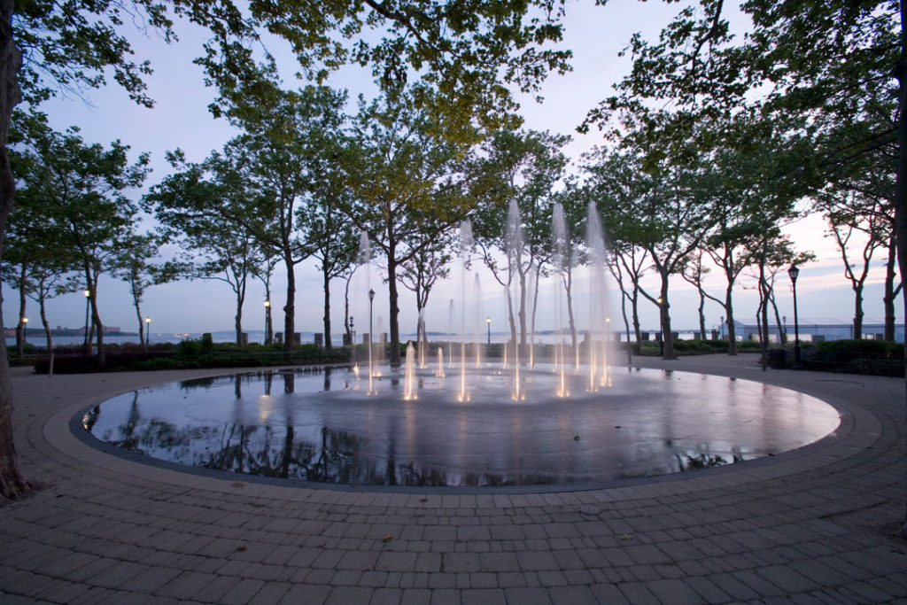 An evening view of the water feature at the Battery Bosque in Battery Park. A spiral of fountains shoot skyward from the ground.  Water pools in a shallow circular area open to passersby to walk through.  Beyond are London plane trees and a view of Governors Island and the New York Harbor.