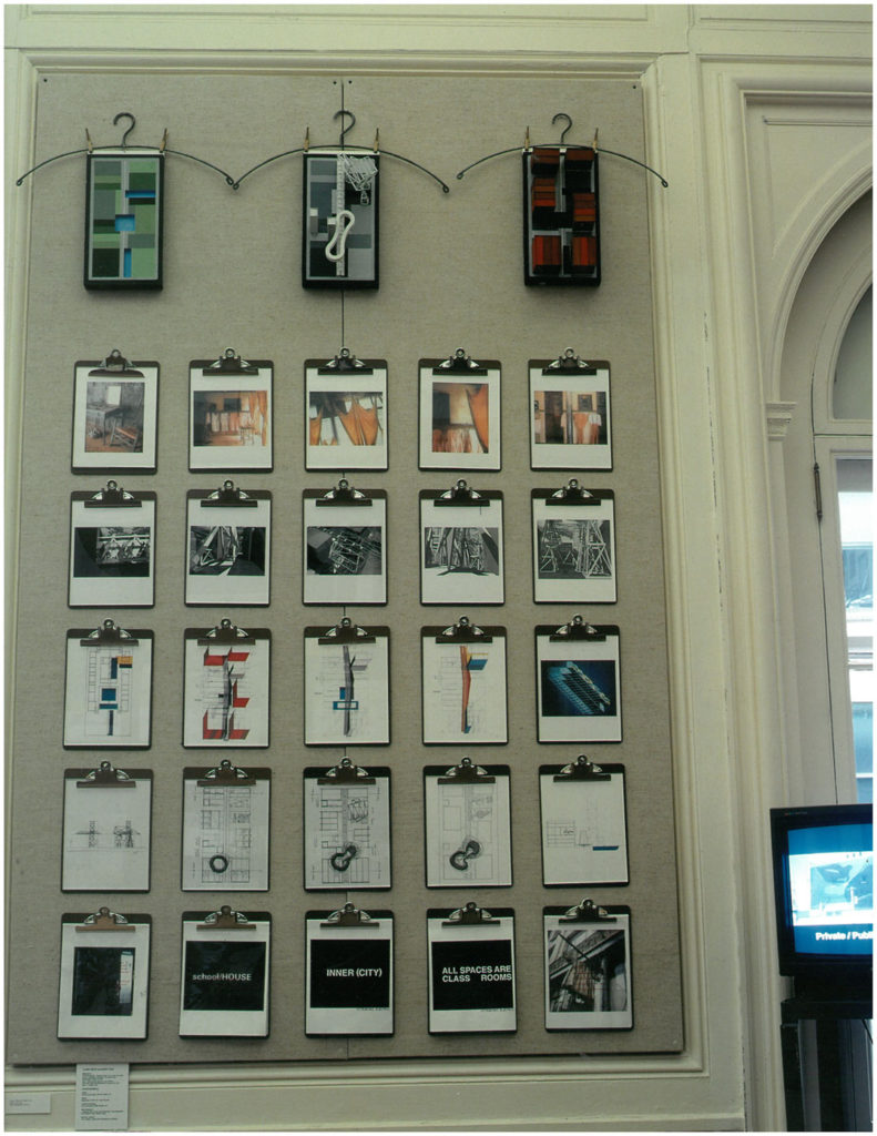 A photograph of the exhibition Beg Borrow Steal.  Rows of clipboards hand on a wall displaying different architectural drawings, renderings, and graphics.  We see the words “school house,” “inner (city),” and “all spaces are class rooms.”