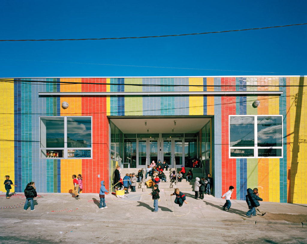 A photograph of the entrance to the Bronx Charter School for the Arts, a one-story building with a tiled exterior made up of brightly colored stripes. Young children gather on the stairs and in the yard to sit, talk with each other, and play jump rope.  It was taken on a clear, bright fall or winter day.