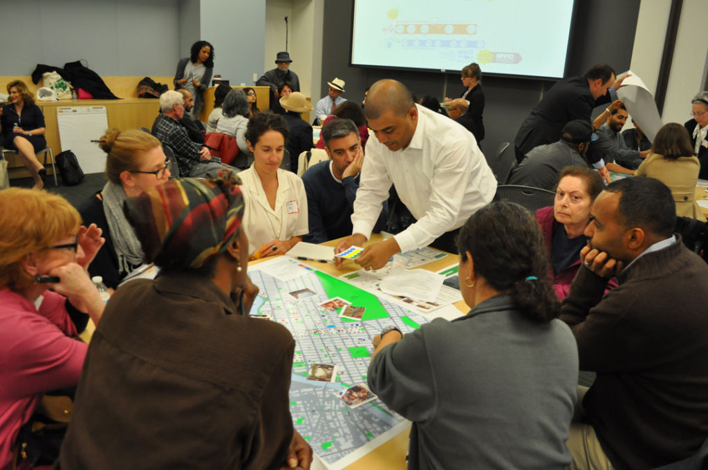 A photo of a community planning meeting organized by Claire Weisz and WXY architecture and urban design in East Harlem.  Groups of adults of different ages and ethnicities gather together around round tables.  A presentation appears on a screen on the back wall.  At the nearest table, we look over the shoulders of two group members, as if we are joining their circle.  A man in a dress shirt stands opposite and adds a paper to the others on table.  The group looks focused and thoughtful, concentrating on a map with different areas marked in colors, photographs, and other documents.