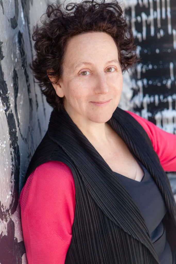 A headshot of Claire Weisz. She leans against a painted concrete wall and smiles welcomingly at the viewer.