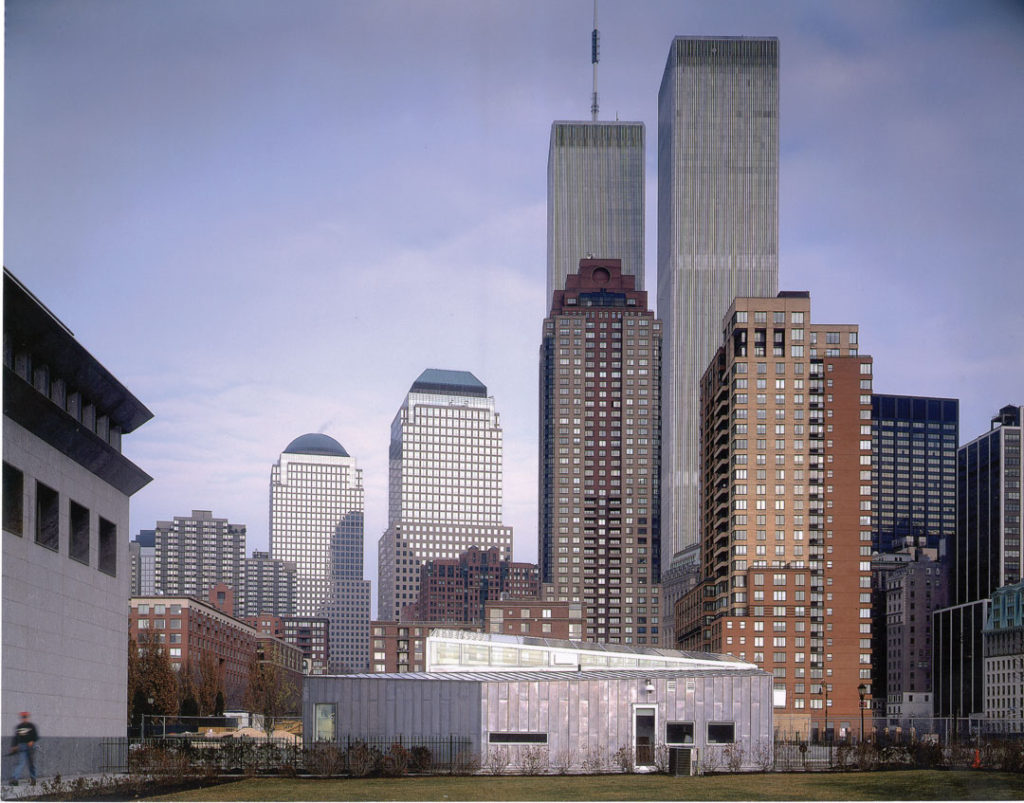 A photograph of a long view of the Museum of Jewish Heritage Visitors Center designed by WXY.  Across a grass lawn is a short, concrete building with a glass, angled roof.  The building sits below towers that make up the Lower Manhattan skyline, including the former World Trade Center.