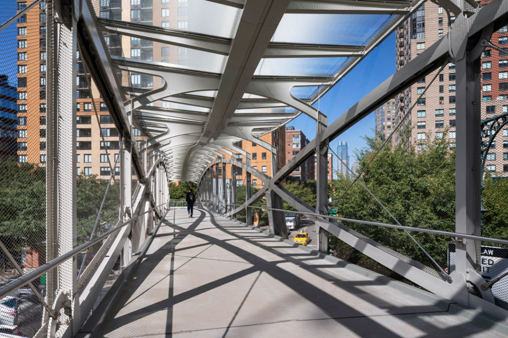 A photograph of the West Thames Bridge, designed by Claire Weisz and WXY architecture and urban design.  The bridge rises diagonally across two intersections of traffic.  It is made of metal and glass and has a small footprint.  Its interior is open, airy, and light and has a design that feels both classic and modern.