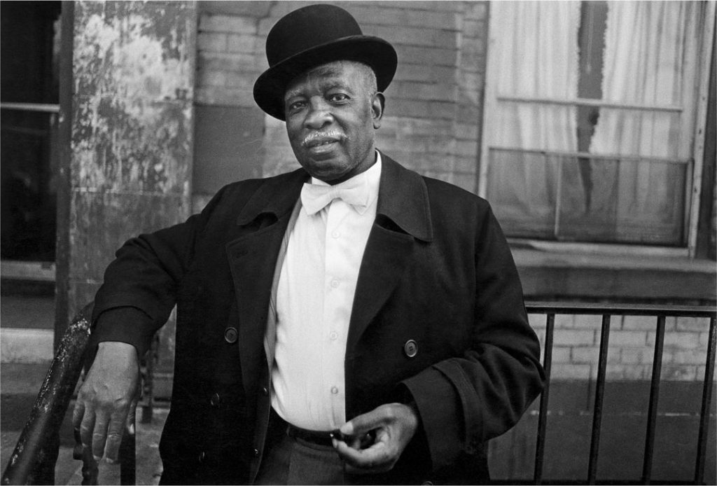 A black and white photograph of a distinguished older Black man clad in a bow tie, dress shirt, overcoat, and bowler hat. He poses outside a brick apartment building, one arm casually draped against a rail. He looks at the camera head-on with strength, dignity, and elegance.