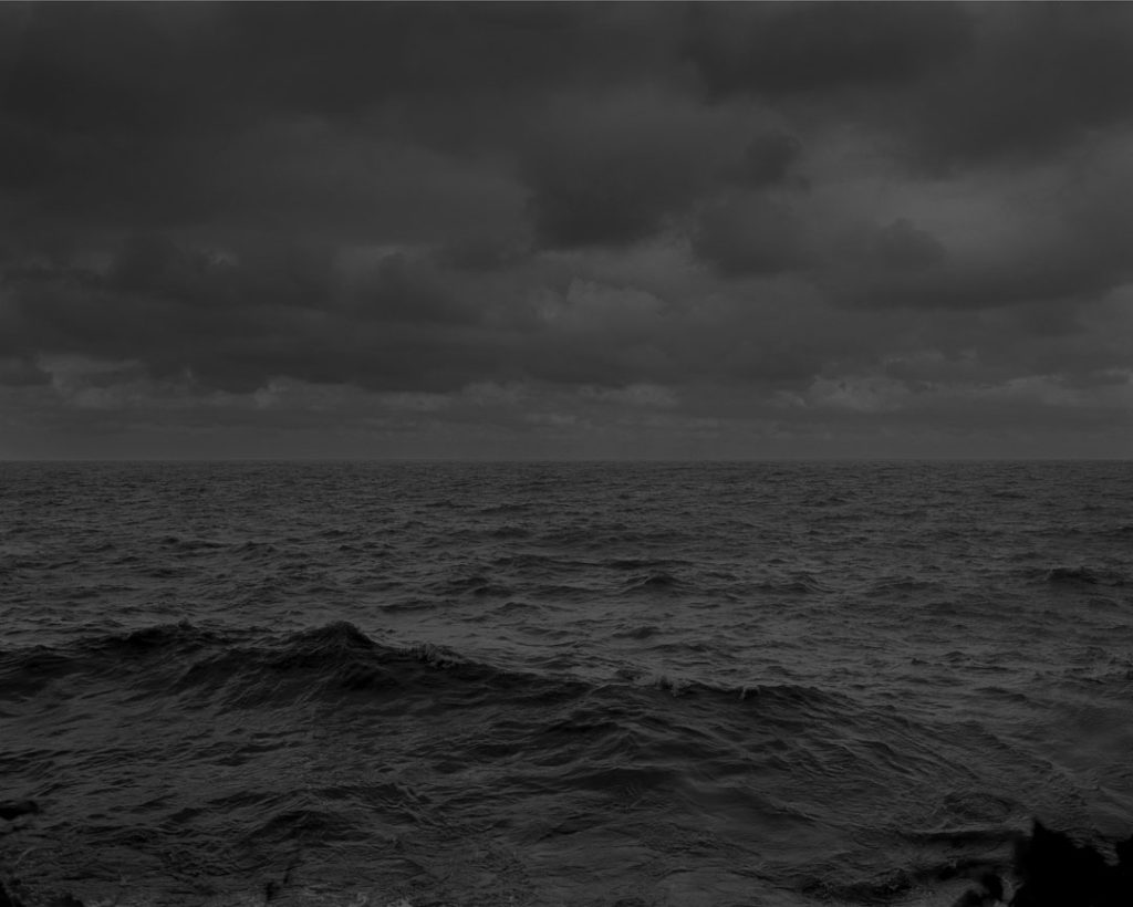 A black and white photograph shot in low light, depicting a large body of water roiling in waves. Overhead are layers of stormy clouds.