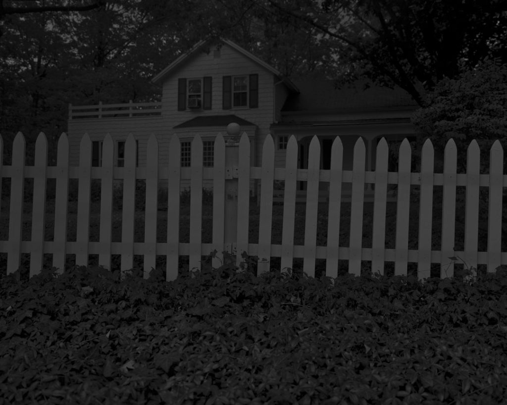 A black and white photograph by Dawoud Bey. Shot in low light, it depicts the a farmhouse, shot from outside its yard which is enclosed by a picket fence.