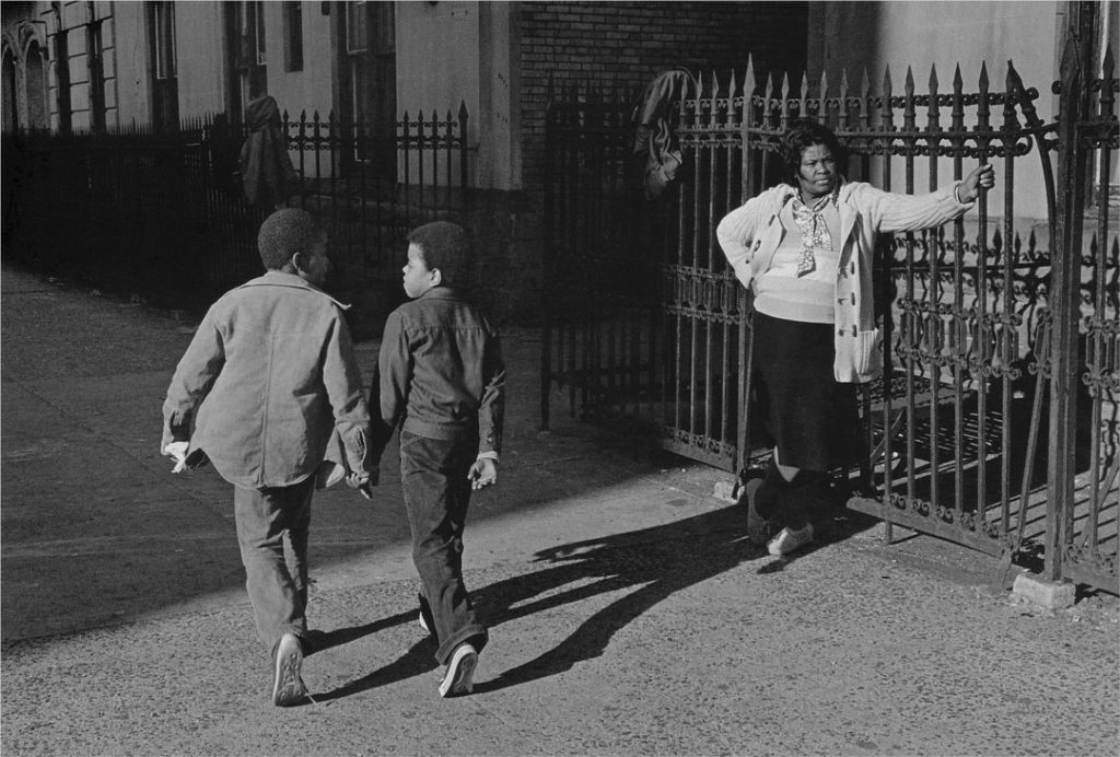 A black and white photograph by Dawoud Bey of a sunny city street. A Black woman wearing a sweater and scarf rests against a wrought iron fence, one hand extended to hold a rail, the other rested on her hip. Her gazes goes beyond the frame, looking down the street as if watching for someone to arrive. Two young boys walk along the sidewalk in the opposite direction, talking animatedly to each other.