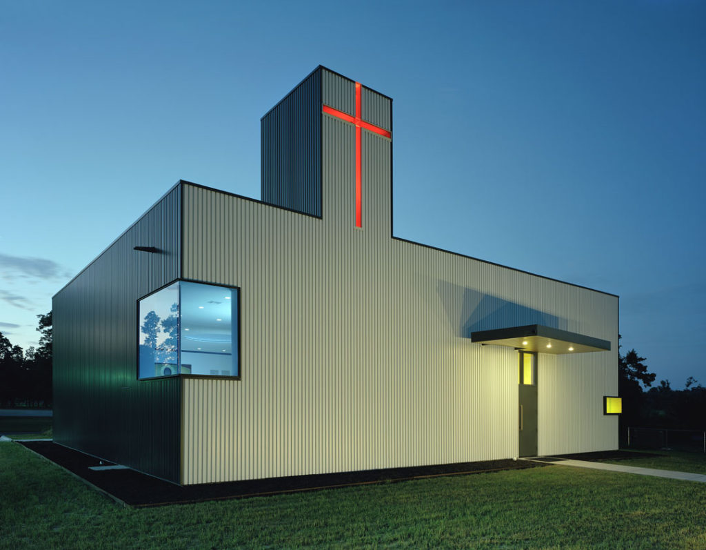 A twilight shot of St. Nicholas Eastern Orthodox Church, designed by Marlon Blackwell Architects.  The rectangular building features a simple gray exterior with a cutout window that wraps around a corner of the building, and a tower that has a cutout of a cross that is lit bright red.