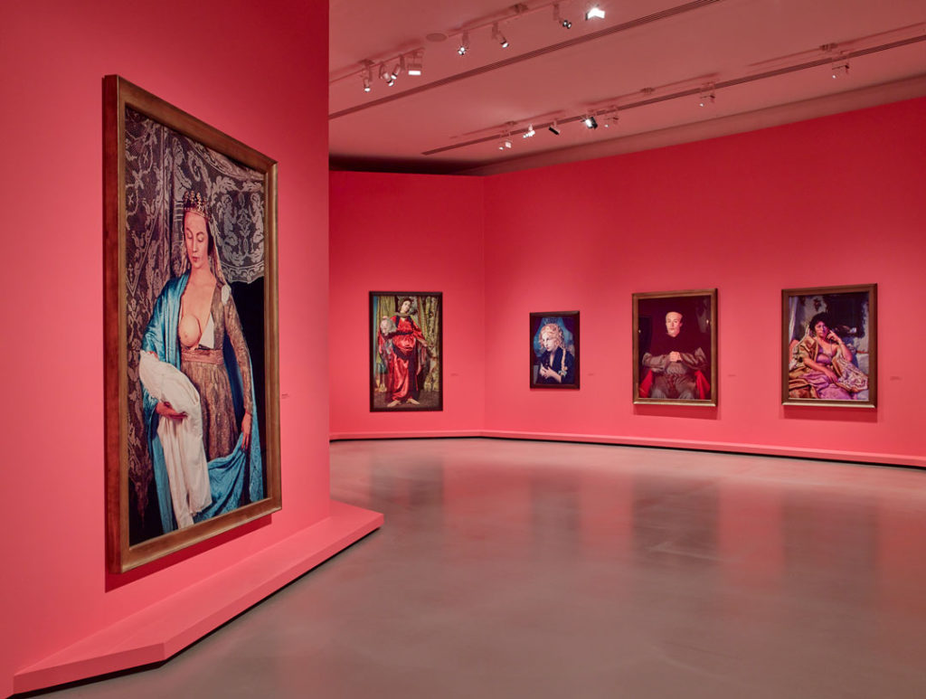 An installation photo of the Cindy Sherman retrospective at the Louis Vuitton Foundation.  This room contains large photographs resembling historic paintings hung on walls painted a rosy neon color. 
